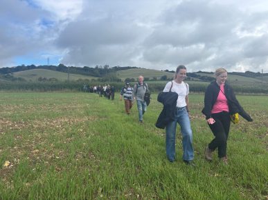A group of people walk along a path across a field. Behind them are open fields on a hillside. The sky above is grey.