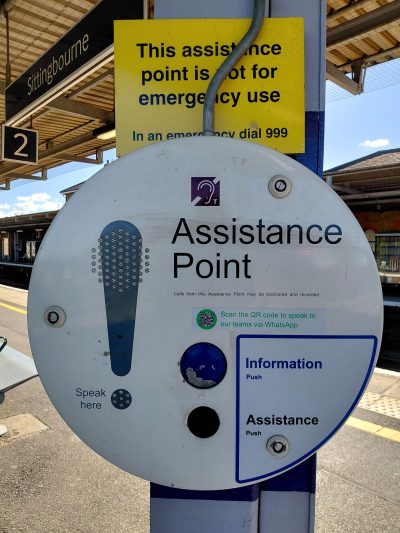 A circular assistance point on a train platform, somewhat like an oversized telephone with a speaker and microphone. There are buttons for Information and Assistance. A notice above explains the assistance point is not for emergency use. A qr code offers connection to support teams via WhatsApp